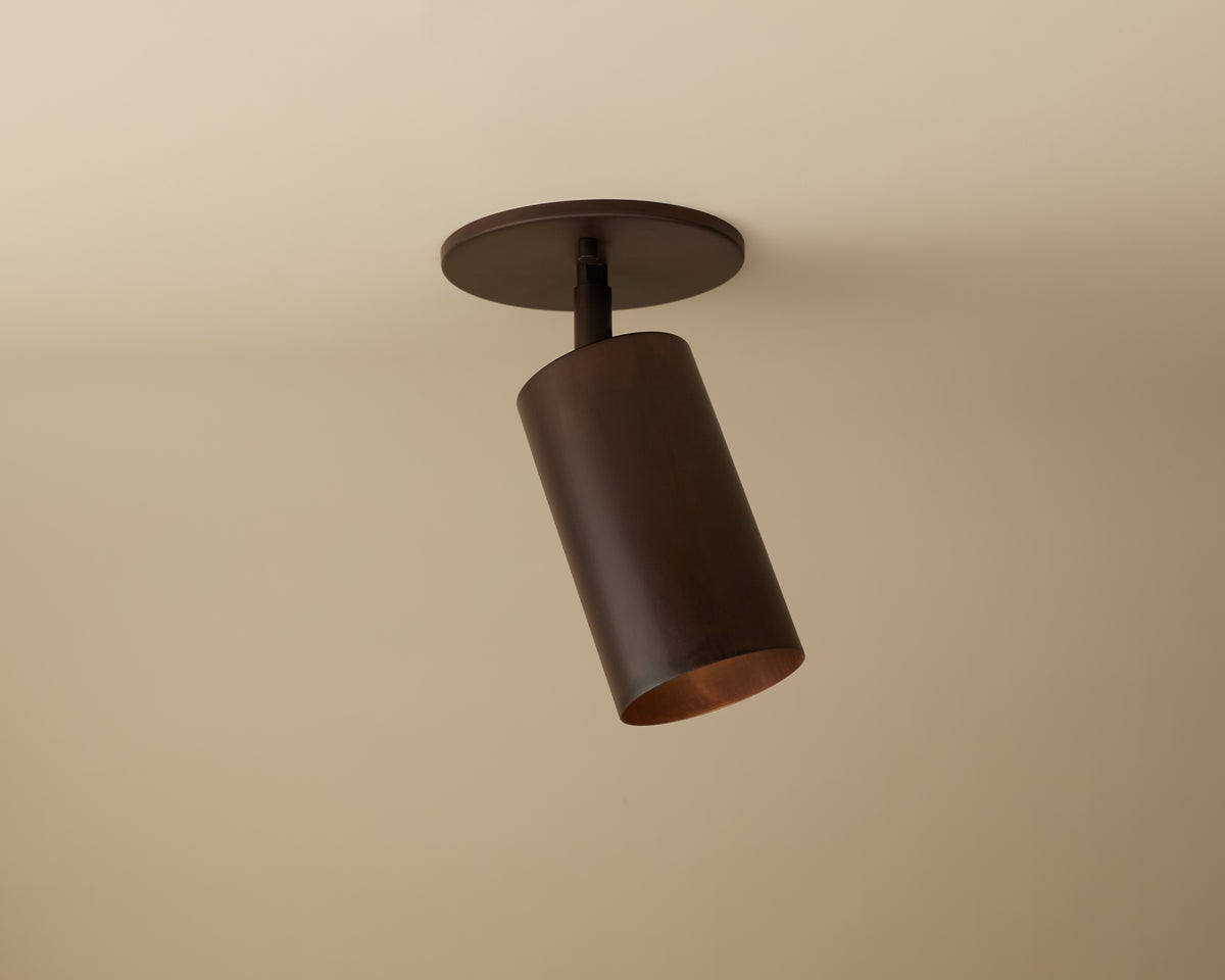 The Long Made Co. Cove Adjustable Spot is a cylindrical, dark brown solid steel downlight with a modern, minimalist design. Mounted on a beige ceiling and angled downward, it’s ideal for custom lighting solutions.