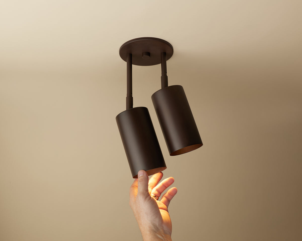 A hand reaches up toward the Long Made Co. Cove Double Drop Spot, a modern dual-cylinder adjustable ceiling downlight with a dark matte finish, mounted on a beige ceiling.