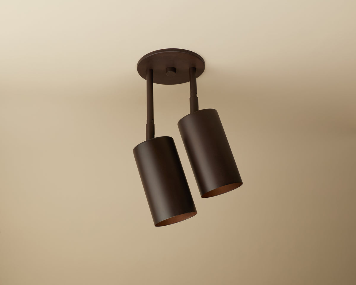 The Long Made Co. Cove Double Drop Spot is a ceiling-mounted downlight with two adjustable, dark brown metal cylindrical shades, providing minimalist, modern lighting against a beige ceiling.