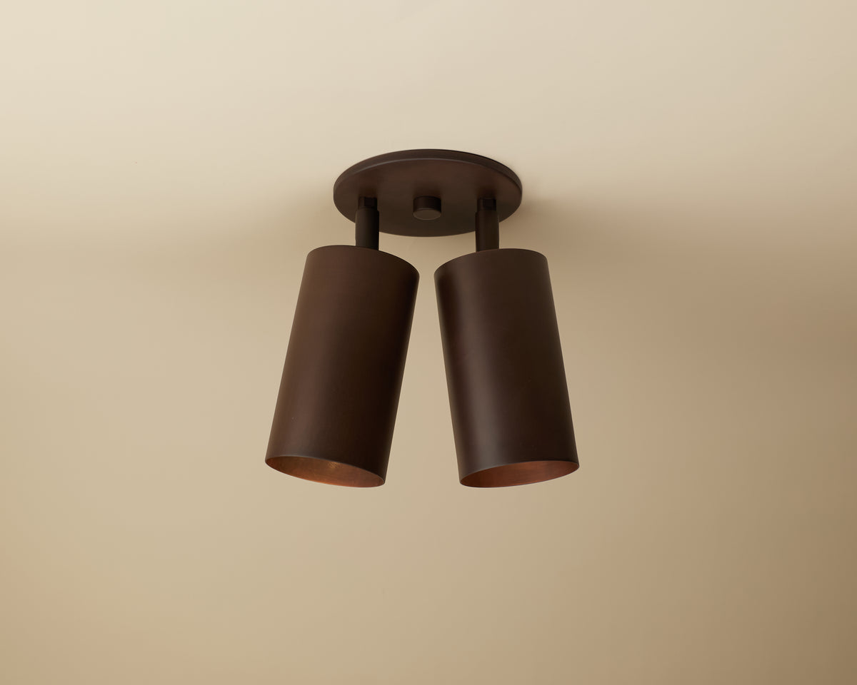 The Cove Double Spot by Long Made Co. features two cylindrical dark brown metal shades on a circular base, angled slightly apart for sleek, minimalist lighting perfect for modern ceilings.