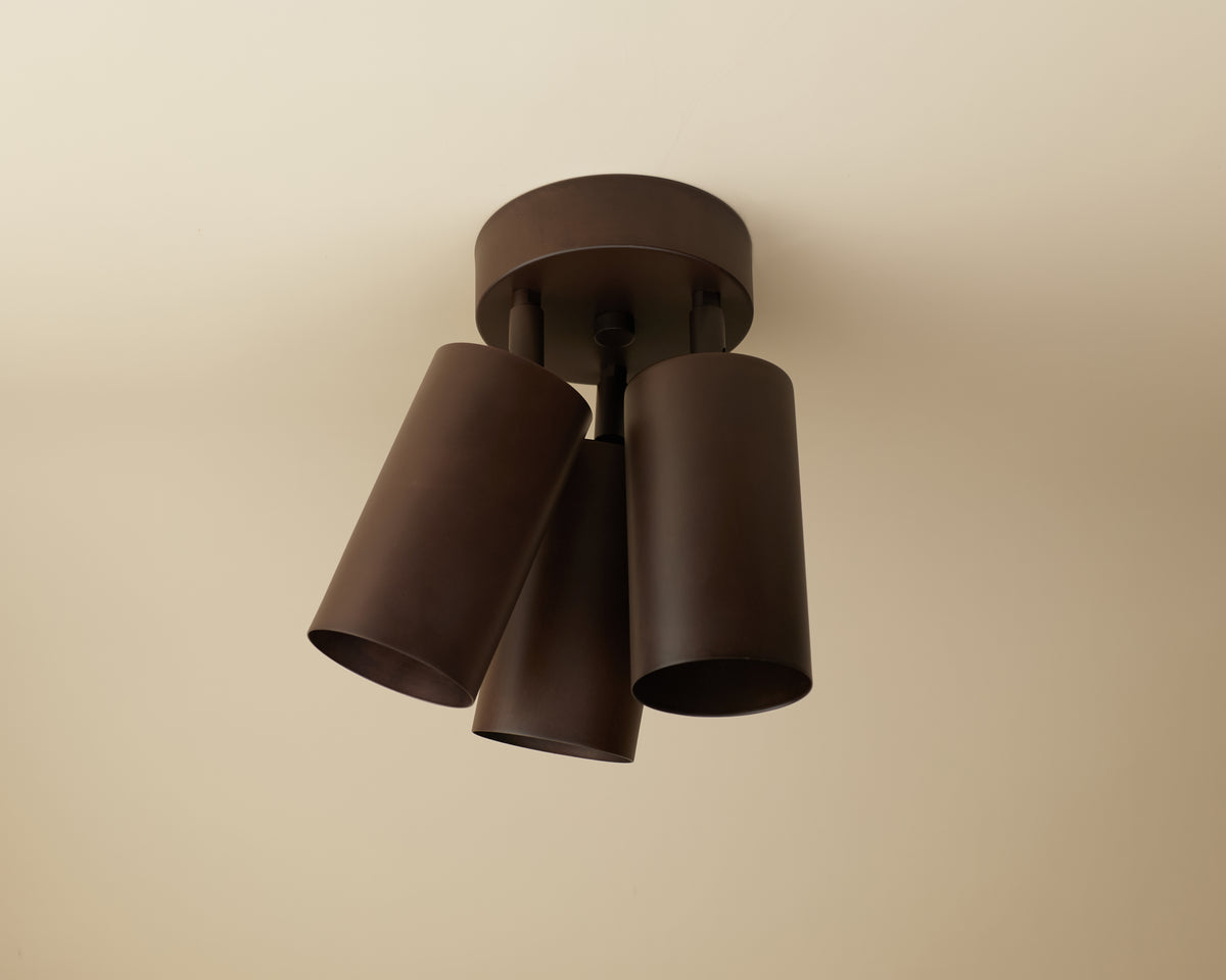 The Cove Double / Triple Spot by Long Made Co. is a ceiling-mounted downlight with three adjustable, dark brown cylindrical metal shades on a circular base—ideal for minimalist lighting.