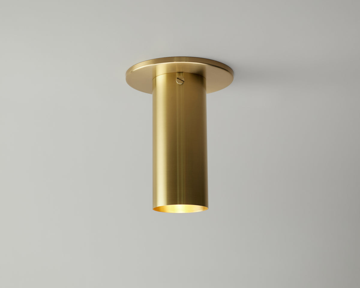 The Brass Cylinder Flush Spot with Long Shade by Long Made Co. is a modern cylindrical brass ceiling light featuring a brushed finish, mounted on a white ceiling and casting a soft, warm downward glow.