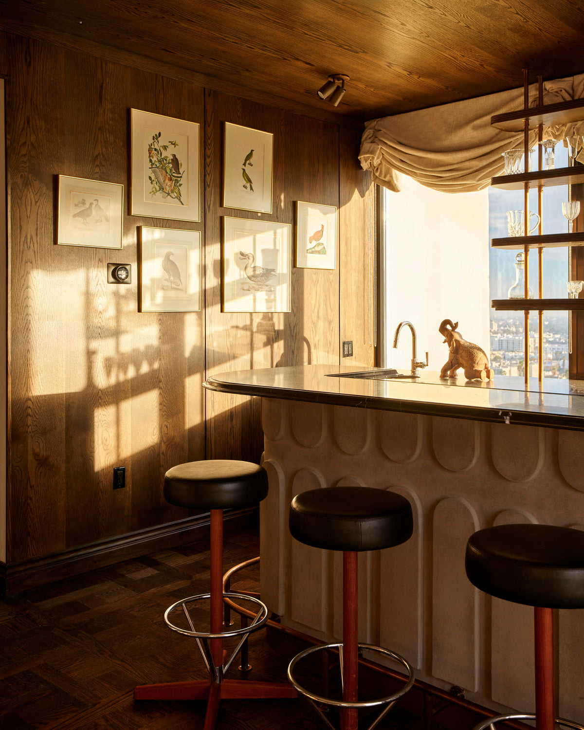 A cozy home bar with a curved counter, two black bar stools, wood-paneled walls with bird art, and sunlit curtains. Overhead, the Long Made Co. Brass Cylinder Triple / Double Spot lighting adds mid-century charm above a small animal sculpture.