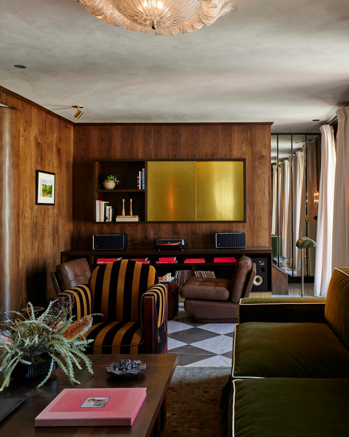 A stylish living room features wood-paneled walls, an olive green sofa, a striped armchair, and a coffee table with books. The Long Made Co. Brass Cylinder Adjustable Spot – Side Mount adds elegance above the gold-accented wall shelf.