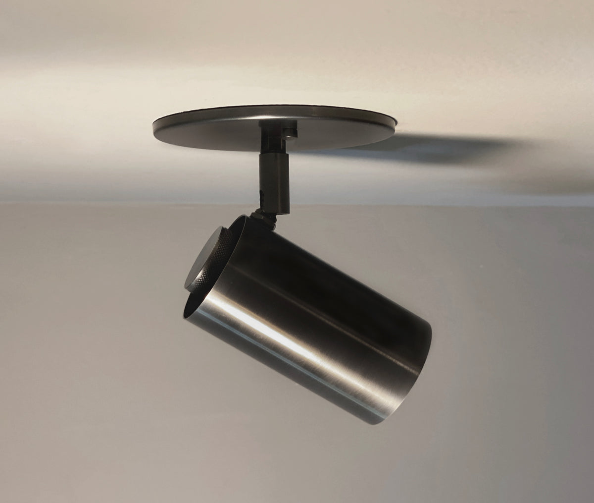 The Brass Cylinder Adjustable Spot - Side Mount by Long Made Co. is a modern cylindrical fixture in black, mounted on a white ceiling and angled downward to illuminate part of the room.
