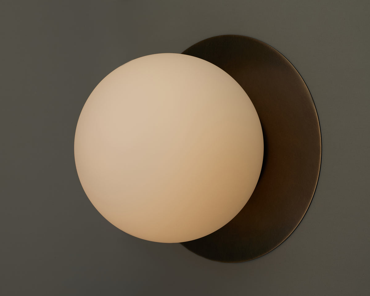 The Sphere Flush by Long Made Co. features a hand-blown, frosted glass shade with a soft glow, mounted on a circular dark metal base against a gray wall—embodying minimal design for a timeless aesthetic.