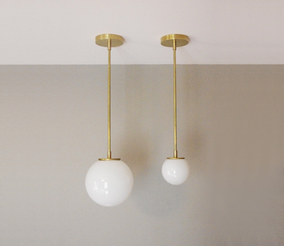 Two Long Made Co. Sphere Solo Drop - 10" solid brass pendant lights with round opal glass shades hang at varying heights in front of a gray wall.