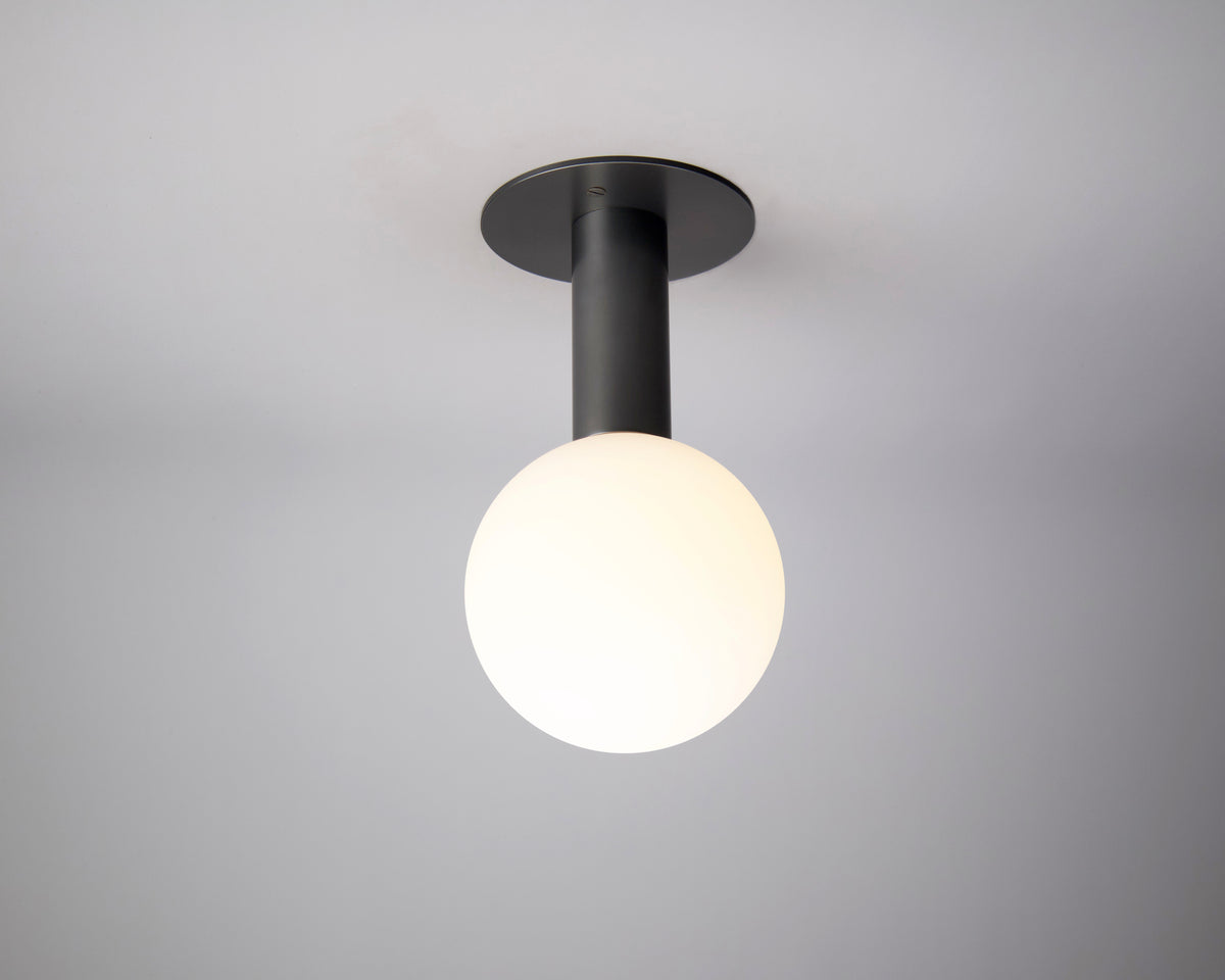 The Sphere Extension by Long Made Co. is a modern handcrafted light with a black base and round matte glass globe, elegantly set in solid brass as a flush mount on a plain white ceiling.