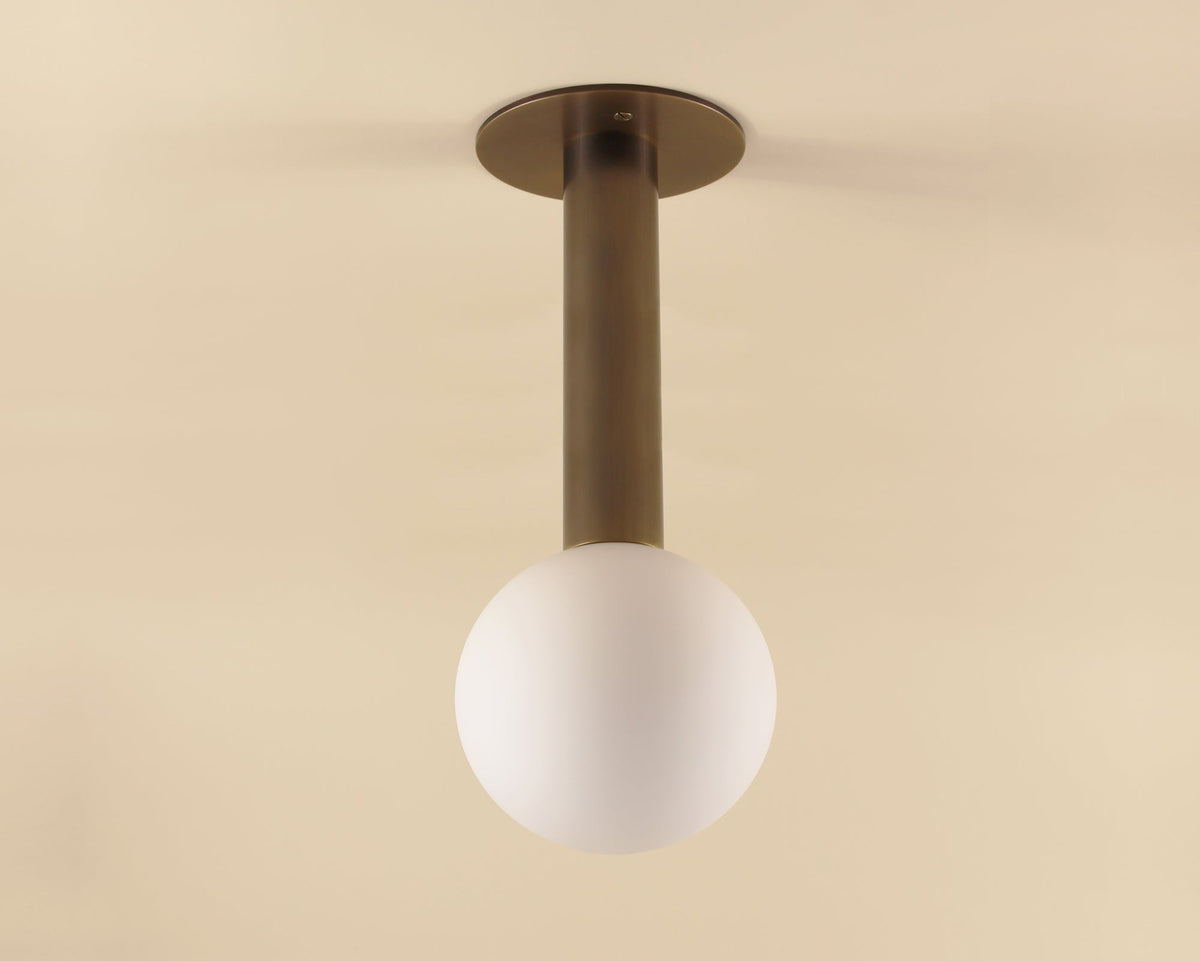The Sphere Drop by Long Made Co. is a modern solid brass flush mount ceiling light with a cylindrical stem and matte glass globe, elegantly installed on a beige ceiling for a custom lighting fixture look.