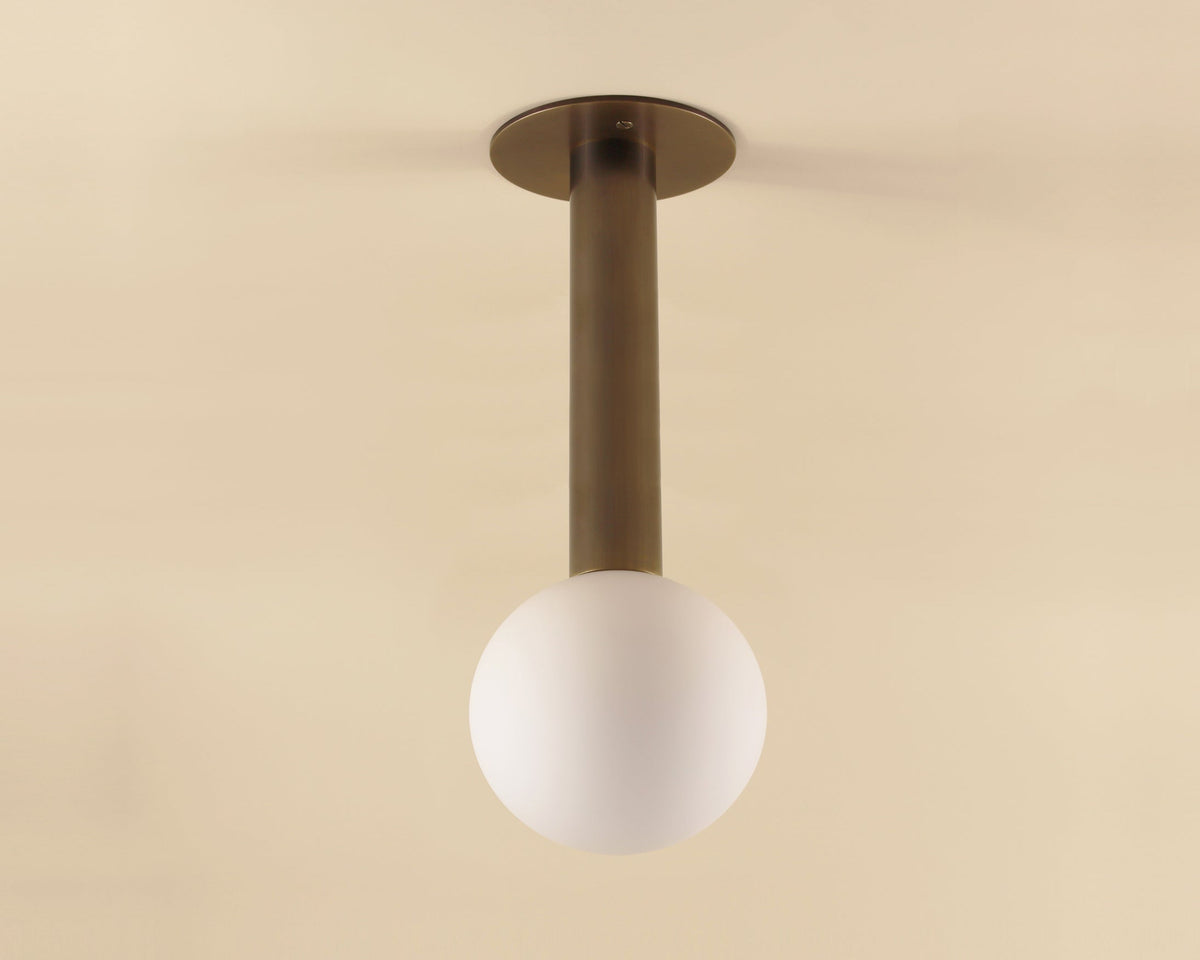 The Sphere Drop flush mount ceiling light by Long Made Co. features a modern brass design with a cylindrical stem and a round matte glass globe hanging downward against a beige background.