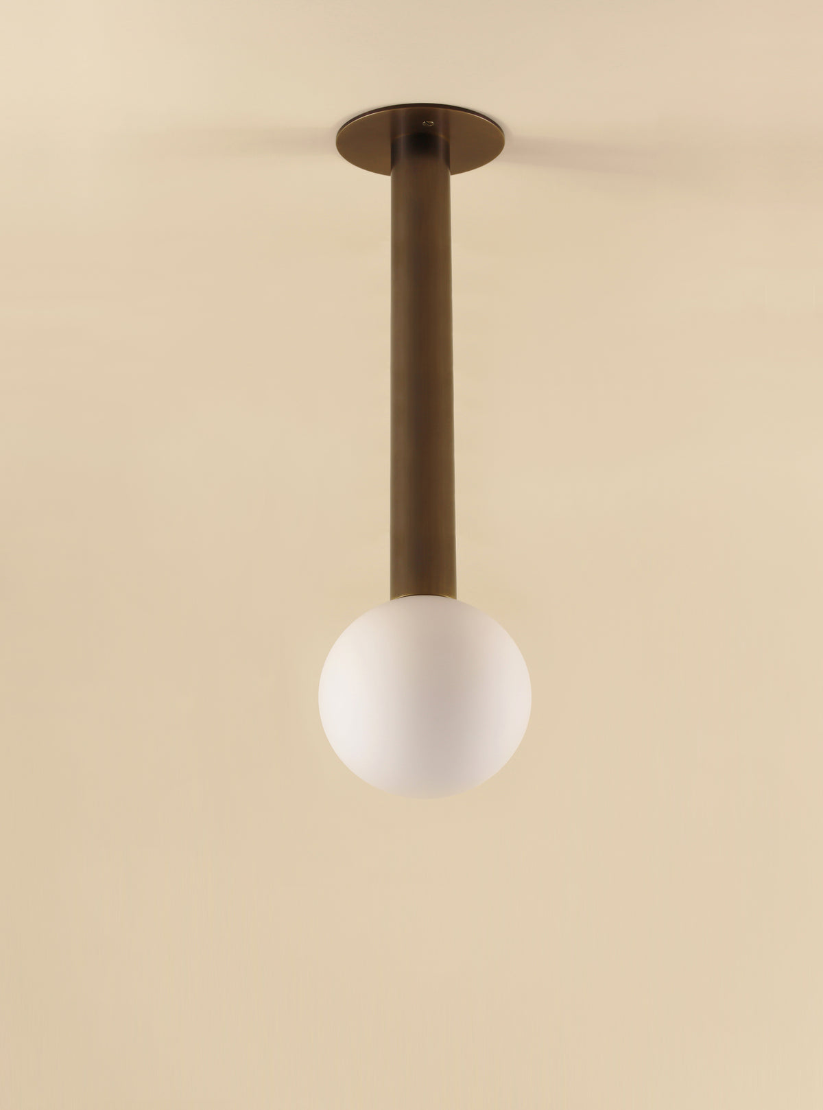 The Sphere Drop by Long Made Co. is a minimalist ceiling light with a matte glass globe and straight, cylindrical solid brass stem, creating an elegant look against a beige ceiling.