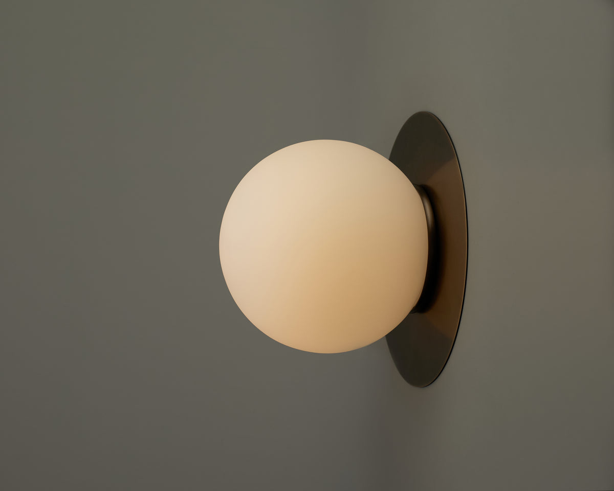 The Sphere Flush by Long Made Co. features a round, hand blown glass shade that emits a soft warm glow, mounted on a dark circular base against a plain gray wall, creating a minimal design statement.