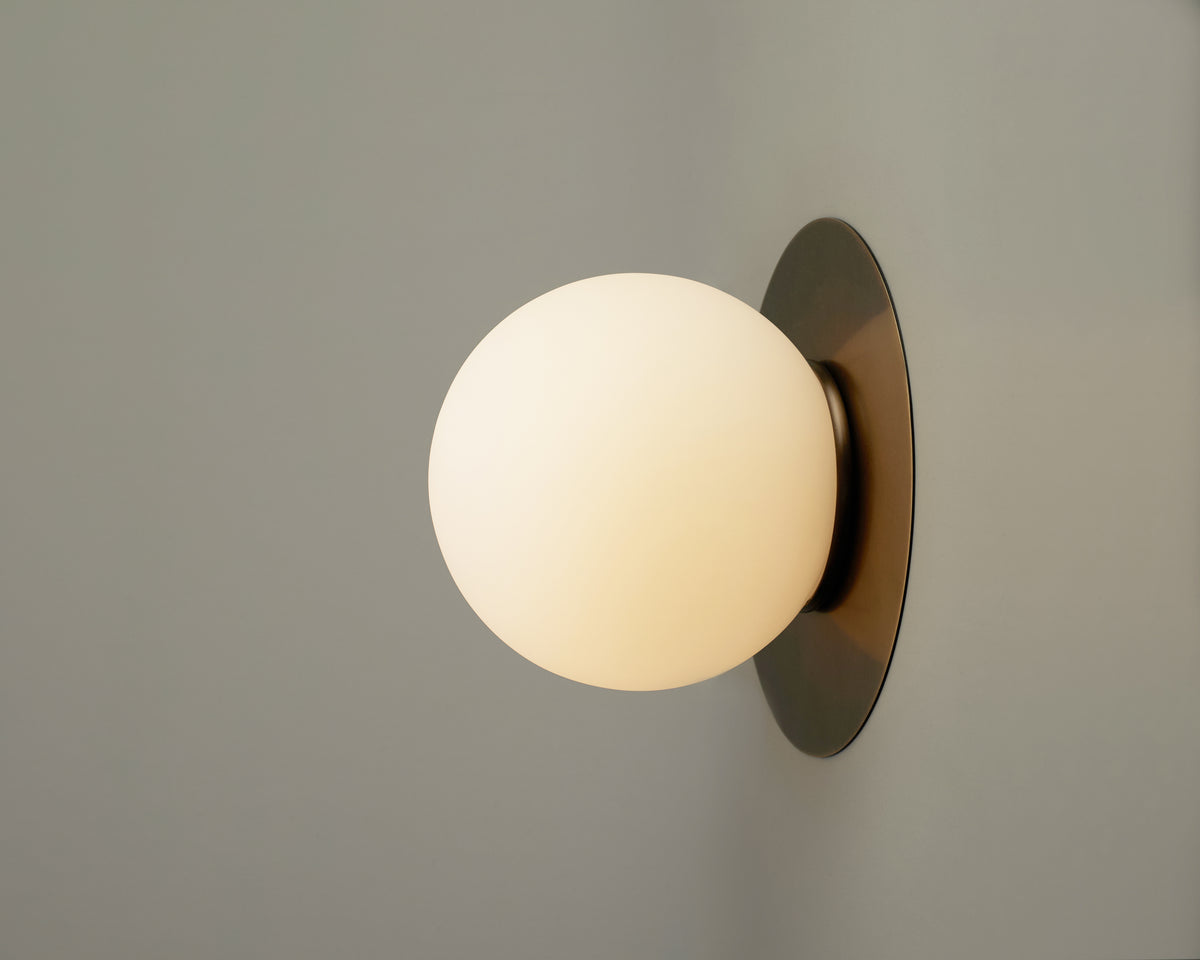 The Sphere Flush by Long Made Co. features a minimal design with a round, frosted hand-blown glass shade and a circular brass base, casting a warm light against a plain light gray wall.