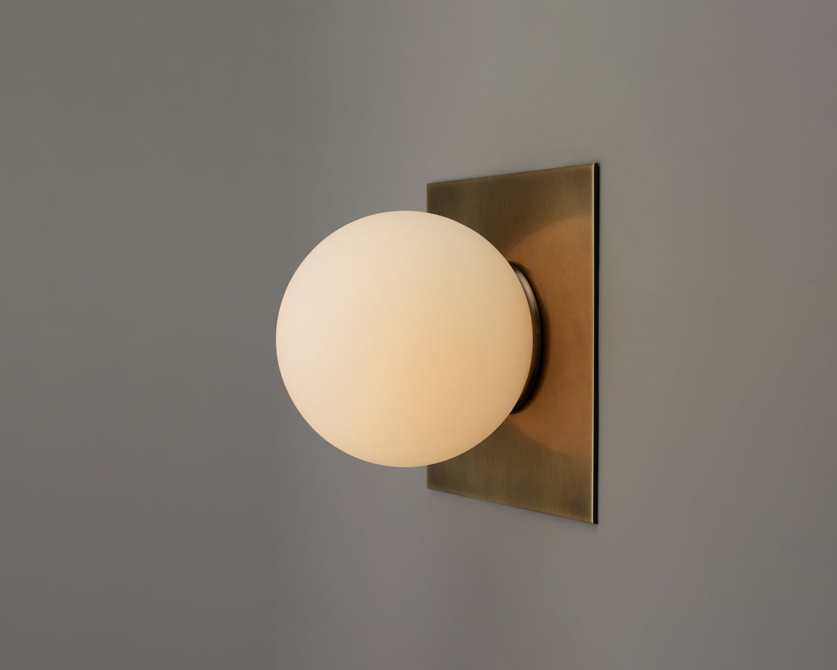 The Sphere Flush by Long Made Co. features a round, hand blown glass shade that casts a warm glow from its rectangular brass plate mount, standing out against a plain gray wall and showcasing the essence of minimal design in this elegant brass wall sconce.