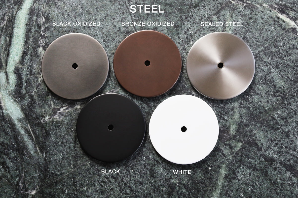 Five round Brass & Steel Finish Samples by Long Made Co. rest on green marble, displaying handcrafted finishes—black oxidized, bronze oxidized, sealed steel, black, and white. Each features a small center hole and unique surface texture.