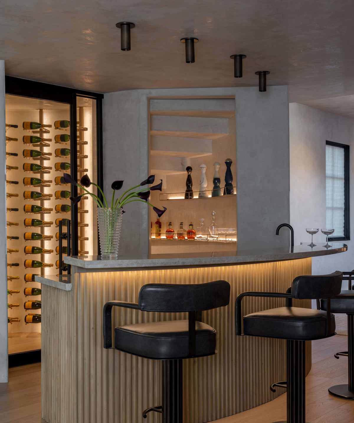 A modern bar with a large island featuring a marble countertop, three black barstools and a decorative vase. Neutral tones prevail, complemented by under-cabinet lighting and Long Made Co.'s Brass Cylinder Flush Spot fixtures above the island.