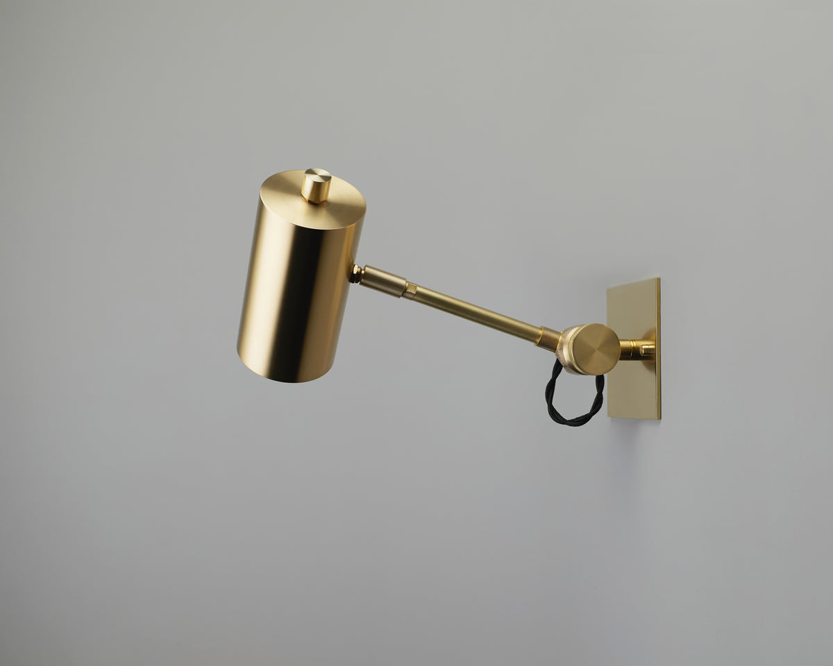 The Task Articulating Wall Lamp Petite by Long Made Co. is a modern gold handmade wall lamp featuring a cylindrical shade and articulating arm, attached to a rectangular wall plate with a visible black cord near the mounting. The background is a plain light gray wall.