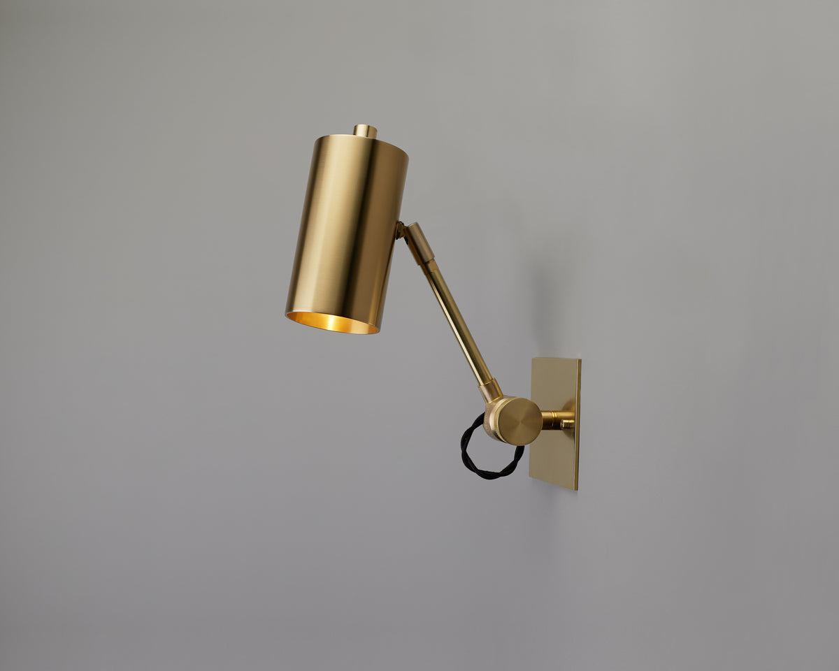A modern, adjustable brass Task Articulating Wall Lamp Petite by Long Made Co. with a cylindrical shade is mounted on a gray wall. Part of our task series lighting, the fixture features a visible black cord looped around the arm for a sleek and functional look.