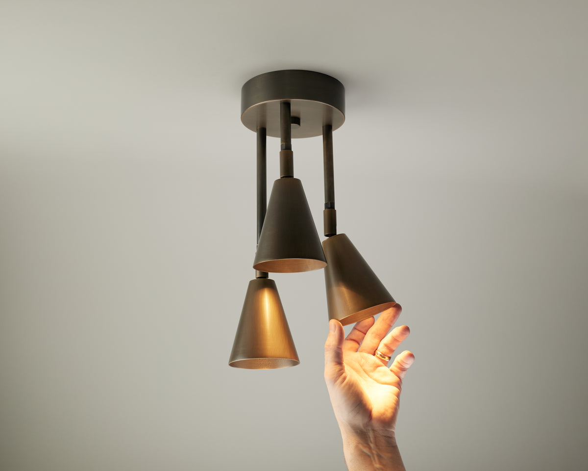 A hand reaches up to adjust one of three cone-shaped Tiel Cascade Drop Spot pendant lights by Long Made Co., attached to a dark fixture, with warm illumination highlighting the hand and lamps—demonstrating the adjustable spot lighting offered by this directional ceiling spot.