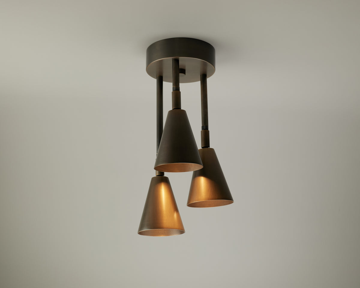 The Tiel Cascade Drop Spot by Long Made Co. is a modern ceiling light fixture featuring three downward-facing, cone-shaped metal shades mounted to a round black base, offering versatile adjustable spot lighting that casts warm light against a plain off-white ceiling.
