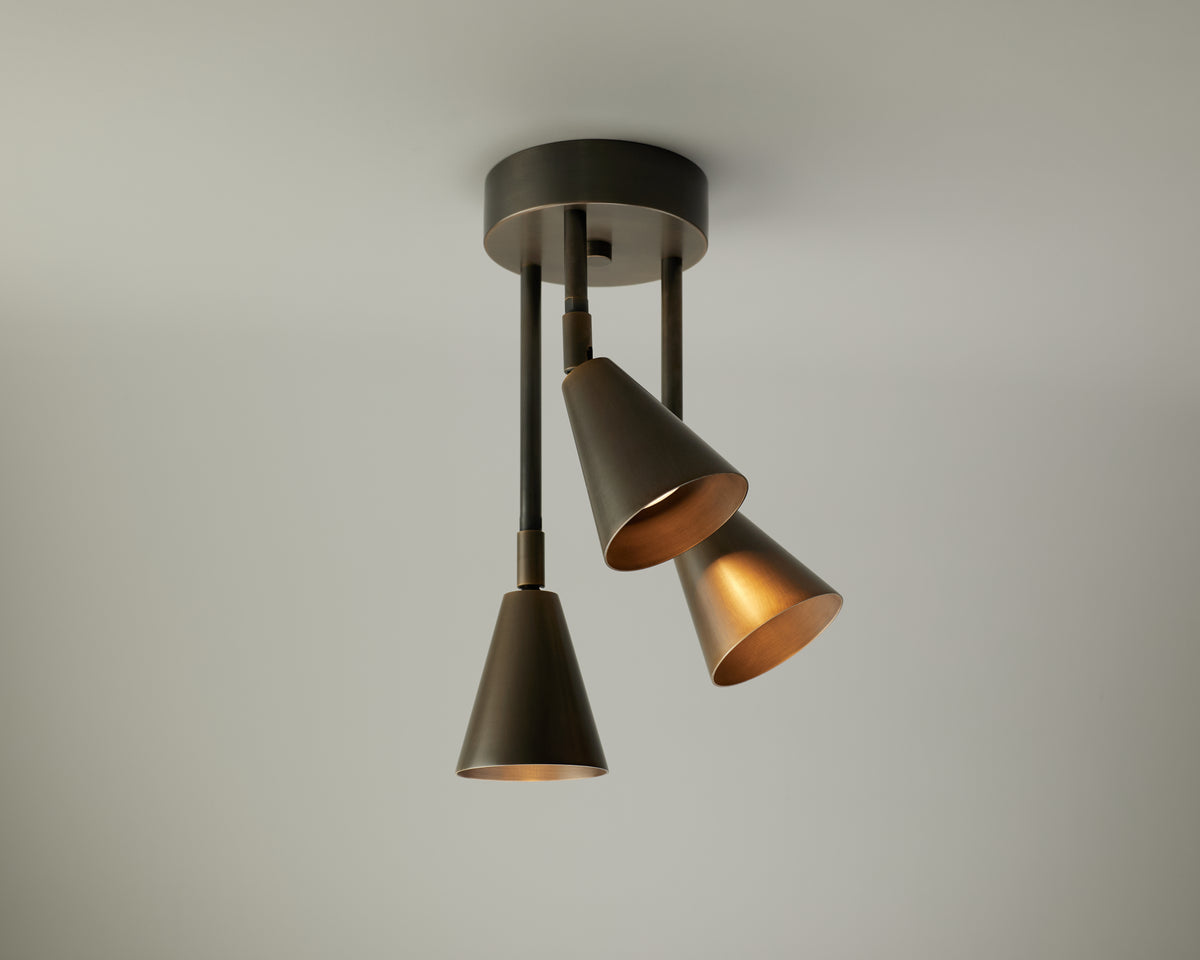 The Tiel Cascade Drop Spot by Long Made Co. is a solid brass ceiling spotlight with three adjustable, bronze-finished, cone-shaped lamps on articulating rods, providing precise directional lighting against a plain light gray background.
