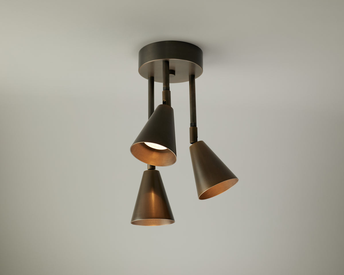 The Tiel Cascade Drop Spot by Long Made Co. is a modern solid brass ceiling spotlight fixture featuring three adjustable cone-shaped metal shades, each oriented in a different direction and mounted on a round base against a plain white ceiling.