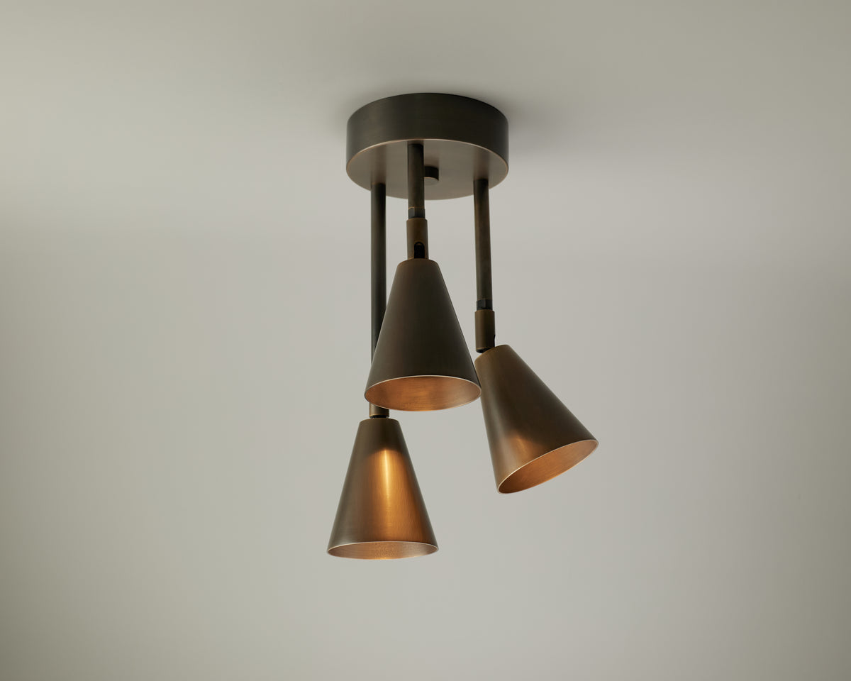 The Tiel Cascade Drop Spot by Long Made Co. is a ceiling-mounted fixture featuring three downward-facing, cone-shaped bronze lamps on sleek cylindrical rods, set against a plain white ceiling. This directional design offers focused illumination, making it perfect for accenting key areas in your space.