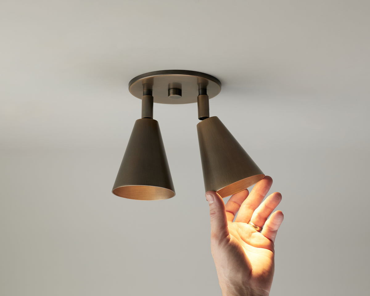 A hand reaches up toward the Tiel Double Spot ceiling fixture by Long Made Co.—a modern, double-cone light inspired by mid-century design—with one cone illuminated and the other unlit, set against a plain white ceiling.