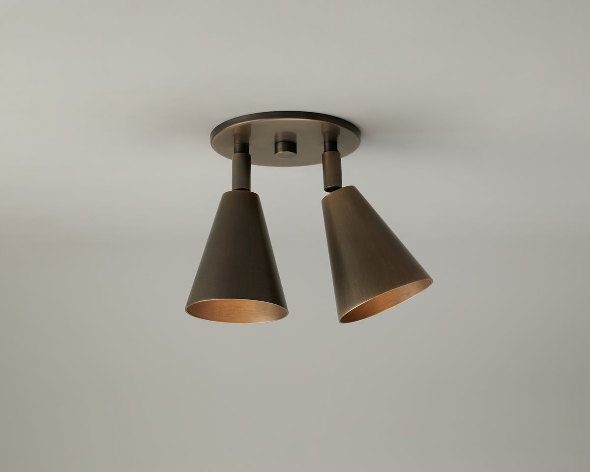 The Tiel Double Spot by Long Made Co. is a modern ceiling light fixture featuring two cone-shaped, bronze-colored metal shades angled downward and mounted on a circular base against a plain white ceiling—a striking example of mid-century lighting that functions as an elegant directional downlight.