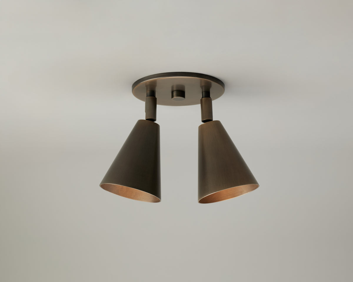 The Tiel Double Spot ceiling light by Long Made Co. features two adjustable, cone-shaped bronze lamps on a round base, providing versatile directional downlighting ideal for functional yet stylish illumination against a plain, light-colored ceiling.