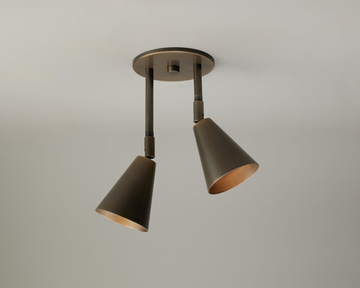 The Tiel Double Drop Spot by Long Made Co. is a modern ceiling light fixture featuring two adjustable, cone-shaped bronze lamps on directional arms, perfect for highlighting features against a plain, light-colored ceiling.