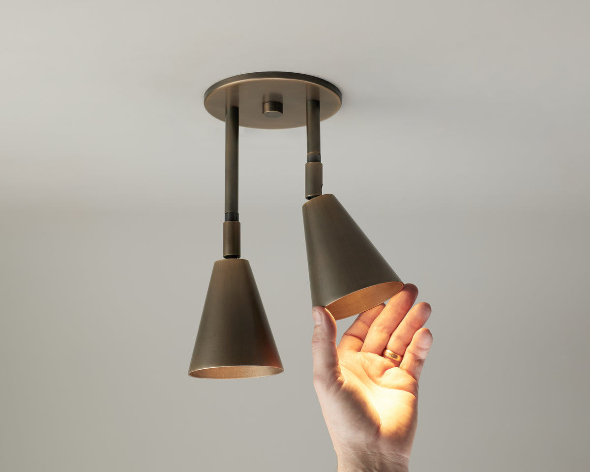 A hand reaches toward the Tiel Double Drop Spot by Long Made Co., a modern solid brass lighting fixture featuring two adjustable cone-shaped lamps—one of which, a directional ceiling spot, is being touched and illuminated.