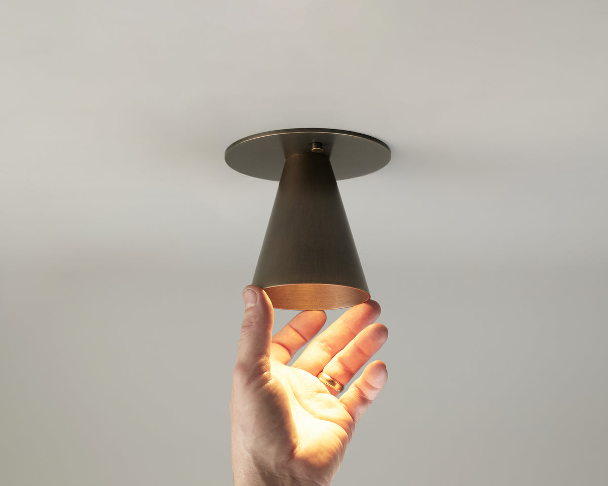 A hand reaches up and touches the modern, cone-shaped Tiel Flush Spot monopoint light fixture by Long Made Co., finished in bronze and turned on, casting a warm glow onto the hand and the surrounding white ceiling.