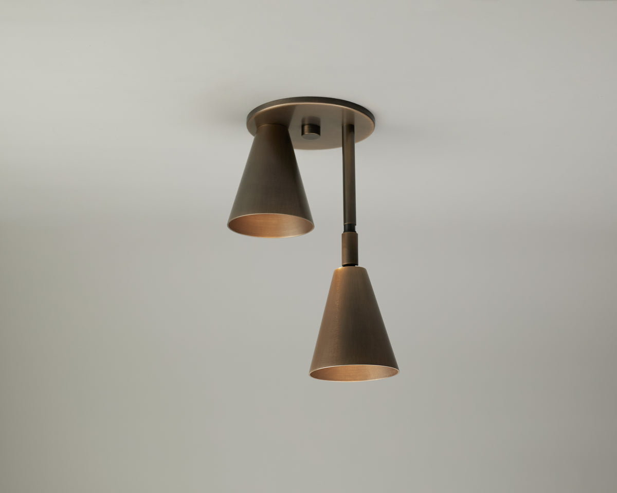 The Tiel Flush / Drop Spot by Long Made Co. is a modern solid brass ceiling light fixture featuring two downward-facing, cone-shaped metal shades, mounted on a round base on a plain white ceiling.