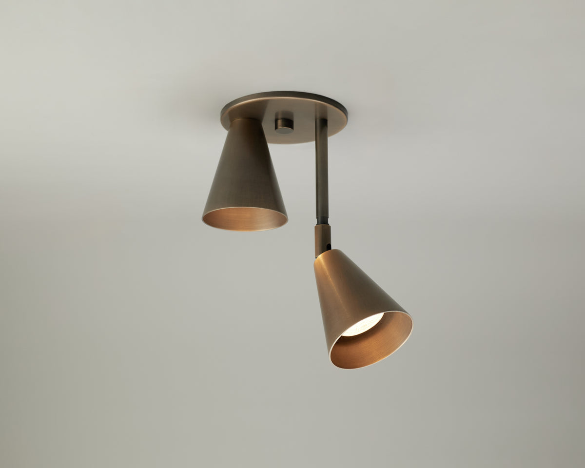The Tiel Flush / Drop Spot by Long Made Co. is a modern ceiling light fixture featuring two cone-shaped bronze lamps oriented in different directions, with one illuminated. Expertly crafted from solid brass and mounted on a plain white ceiling, this elegant and adjustable spot light provides a contemporary lighting solution for sophisticated interiors.