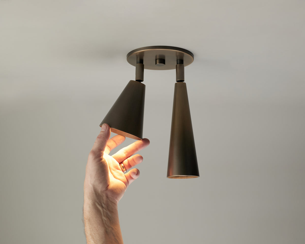 A hand reaches up to touch one of two black, cone-shaped solid brass Long Made Co. Tiel / Pine Double Spot ceiling downlights; one light is on, illuminating the hand, while the other remains off.