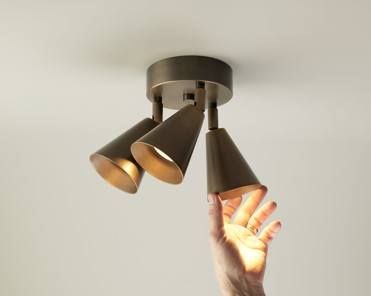 A hand adjusting one of three bronze cone-shaped directional downlights—two illuminated and the third being touched—the Tiel Triple Spot by Long Made Co. is a striking mid-century ceiling light that brings both style and functionality to any space.