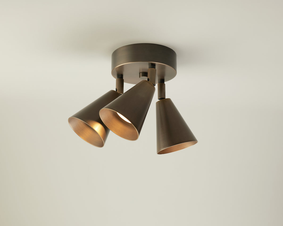 The Tiel Triple Spot by Long Made Co. is a ceiling light fixture featuring three cone-shaped, bronze metal lampshades mounted on a round base and angled in different directions for adjustable task lighting with a mid-century downlight style, shown here against a plain off-white ceiling.