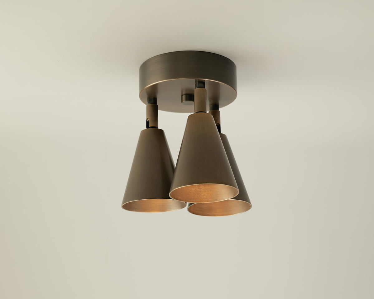 The Tiel Triple Spot by Long Made Co. is a modern ceiling light fixture featuring a round base and three downward-facing, cone-shaped bronze lamps that capture the elegance of mid-century lighting, offering sleek, directional downlight against a plain white ceiling.