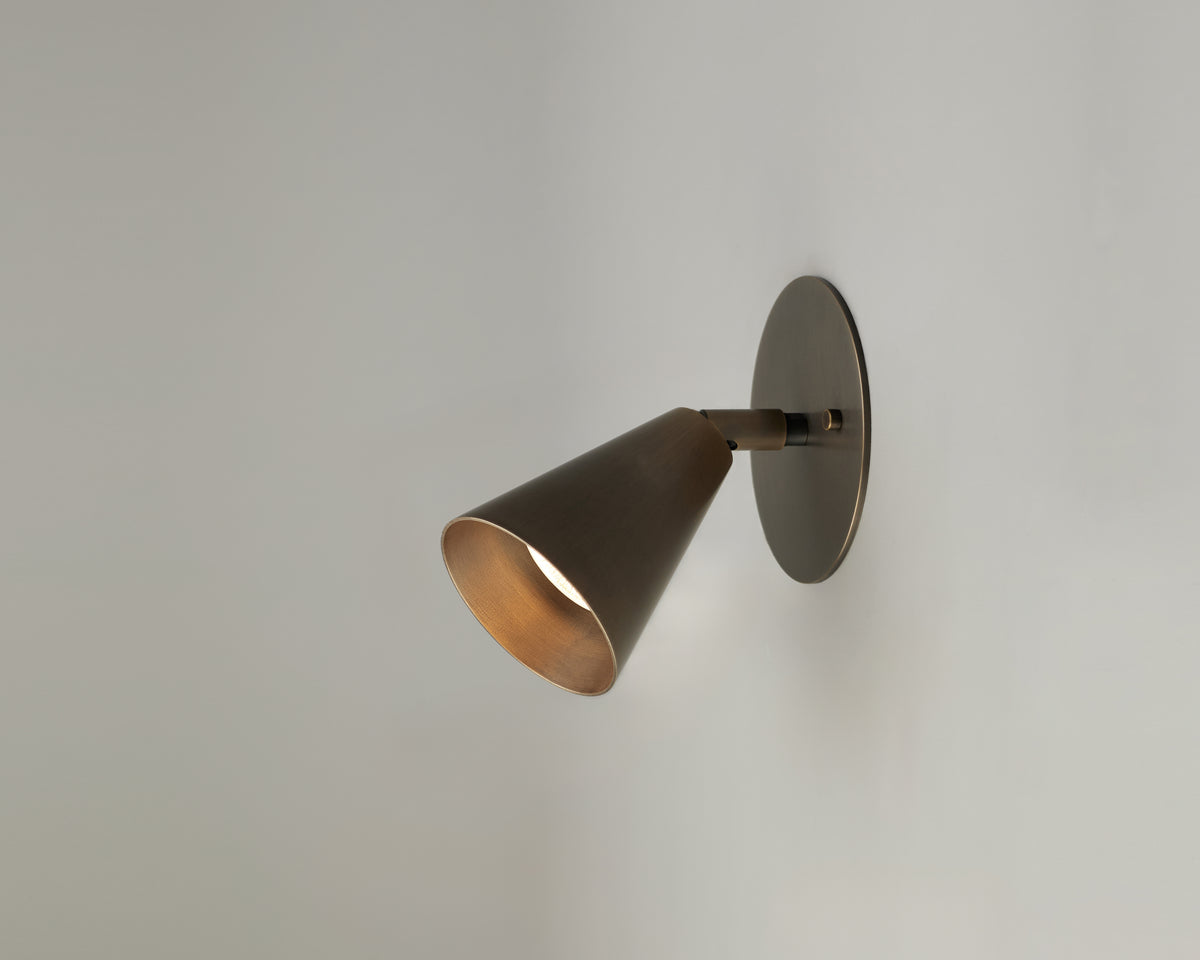 The Tiel Wall Spot by Long Made Co., a modern cone-shaped brass wall sconce, is mounted on a plain light gray wall, casting a soft downward glow.
