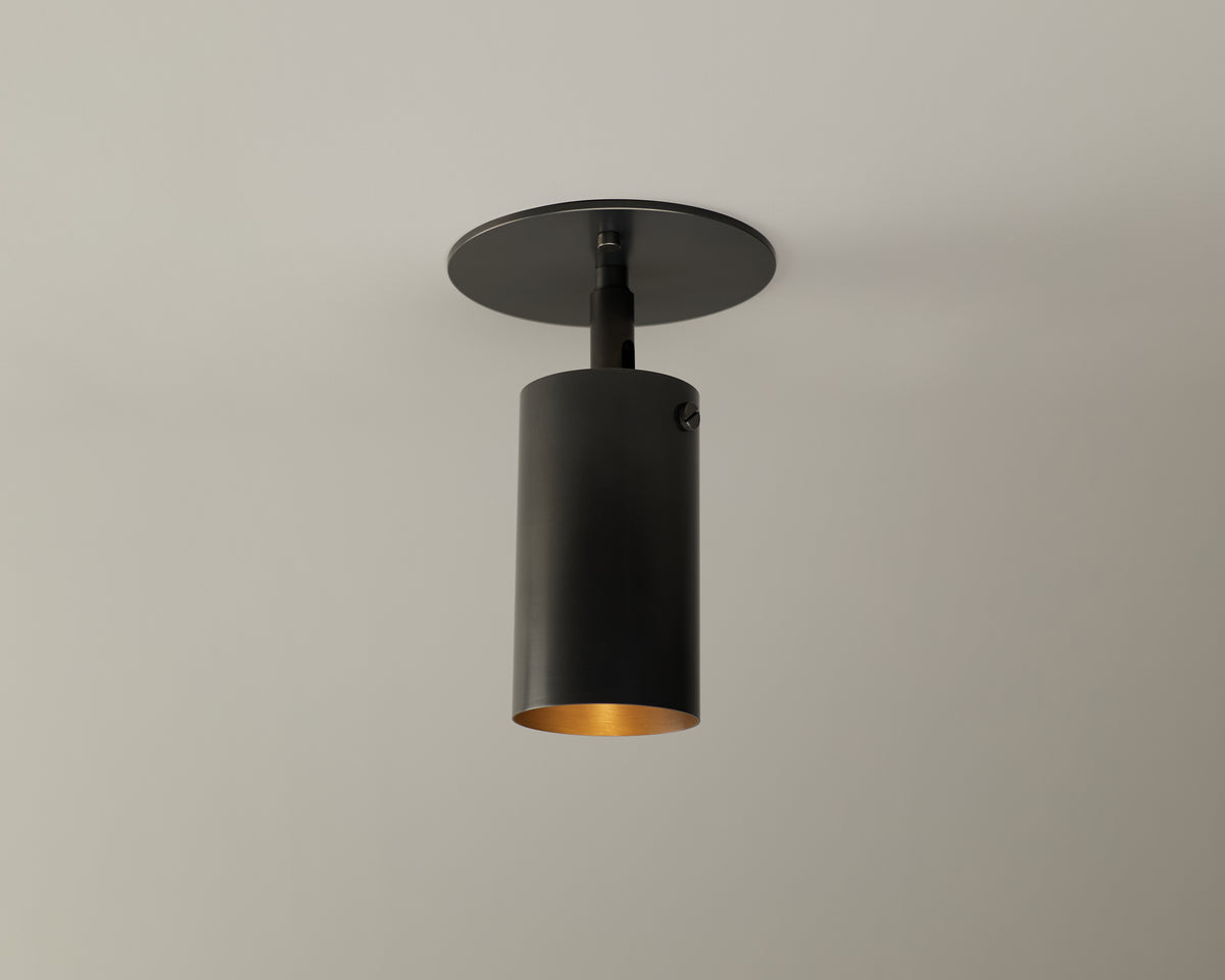 The Long Made Co. Brass Cylinder Adjustable Spot - Top Mount is a modern black cylindrical fixture with a matte finish, set on a round black base against a light ceiling, offering adjustable, downward-focused mid-century modern lighting.