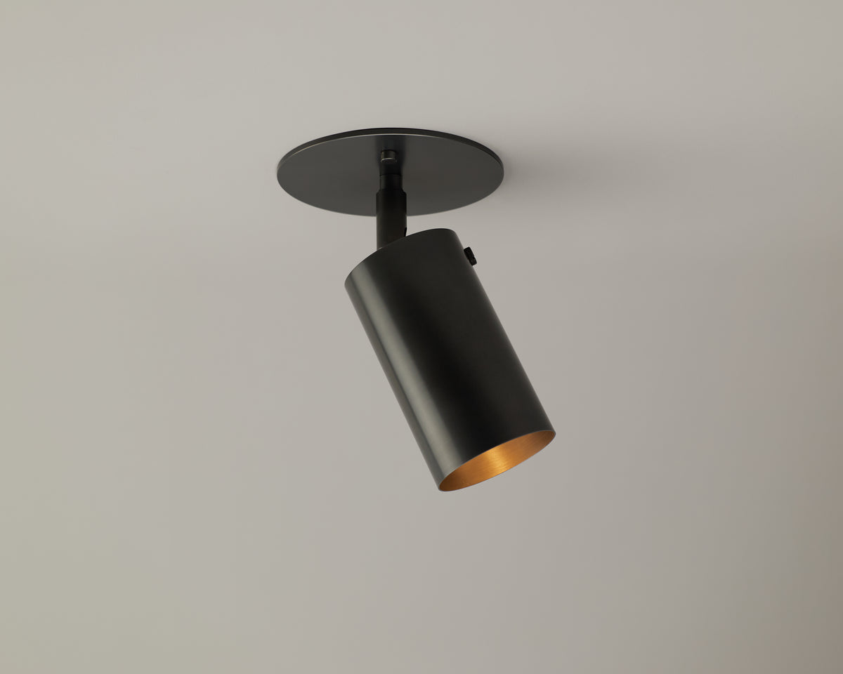 The Brass Cylinder Adjustable Spot - Top Mount by Long Made Co. is a modern cylindrical fixture in brass, mounted on a round base against a white ceiling, angled downward with its interior reflecting warm light.