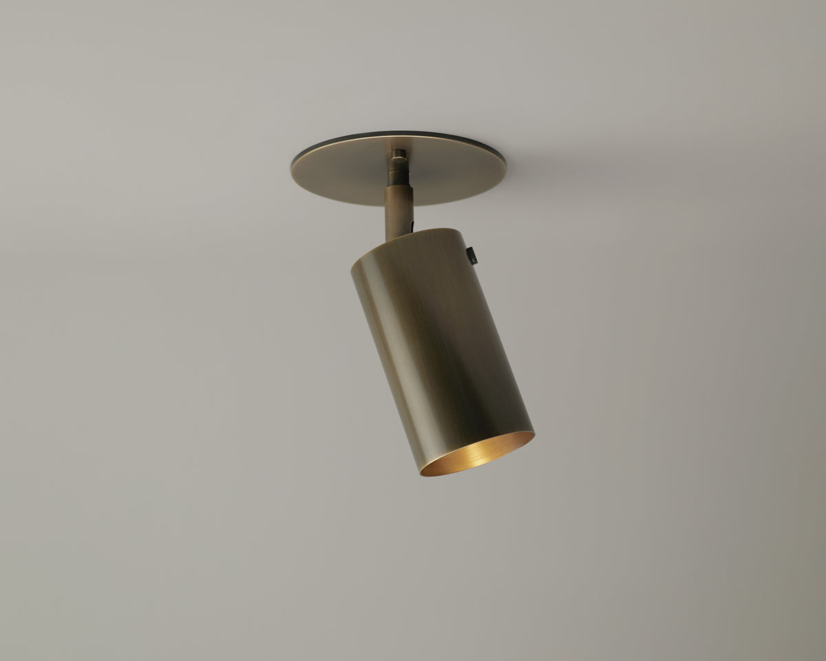 The Long Made Co. Brass Cylinder Adjustable Spot – Top Mount is a cylindrical, brass directional ceiling fixture set on a round base and angled downward against a plain light gray ceiling.