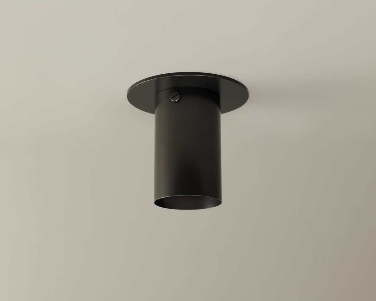 The Brass Cylinder Flush Spot by Long Made Co. is a black cylindrical ceiling light with a round base, mounted on a plain white ceiling, offering clean lines and timeless mid-century modern style.