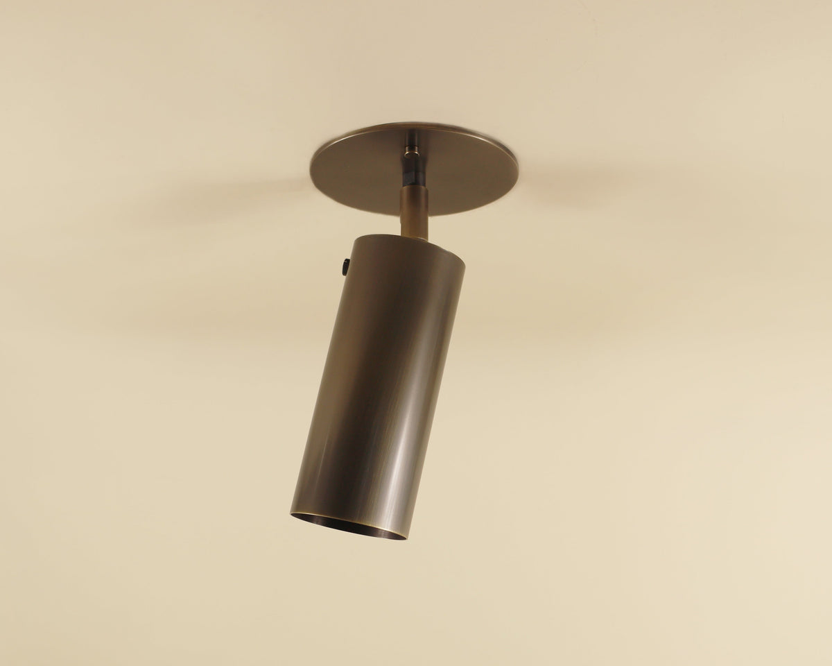 The Brass Cylinder Adjustable Spot - Top Mount w/ Long Shade by Long Made Co. is a sleek, modern brass ceiling light with a cylindrical design, inspired by mid-century style and mounted on a beige ceiling for adjustable directional lighting.