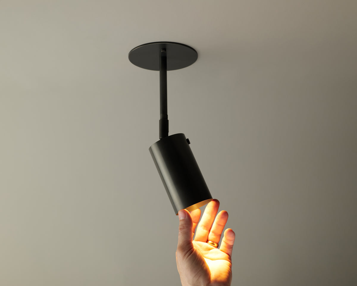 A hand reaches up to touch the light from the Long Made Co. Brass Cylinder Drop Spot - Top Mount, a black directional ceiling fixture with a round base, shown against a plain gray background.
