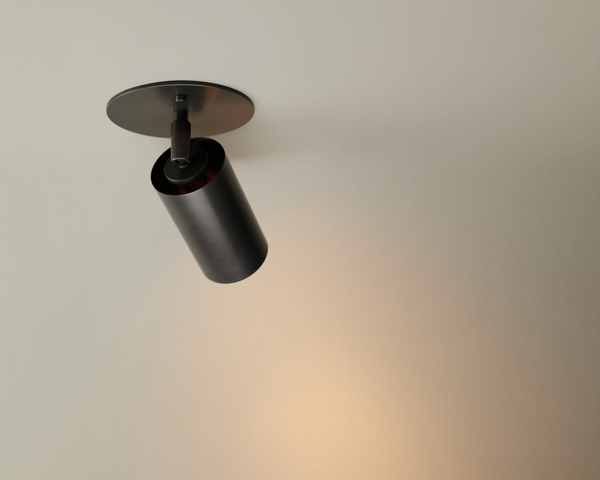 The Brass Cylinder Adjustable Spot - Top Mount by Long Made Co. is a black cylindrical ceiling spotlight that provides warm, focused task lighting and creates a soft glow on the surface below.