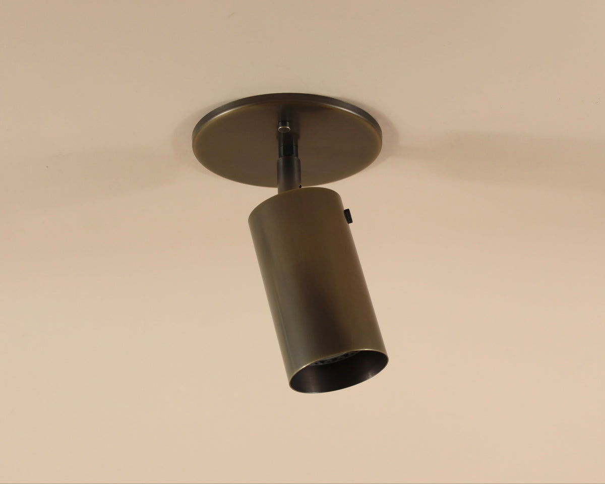 The Brass Cylinder Adjustable Spot - Top Mount by Long Made Co. is a cylindrical brass ceiling downlight with a round base, angled downward on a beige ceiling for a sleek mid-century modern touch.