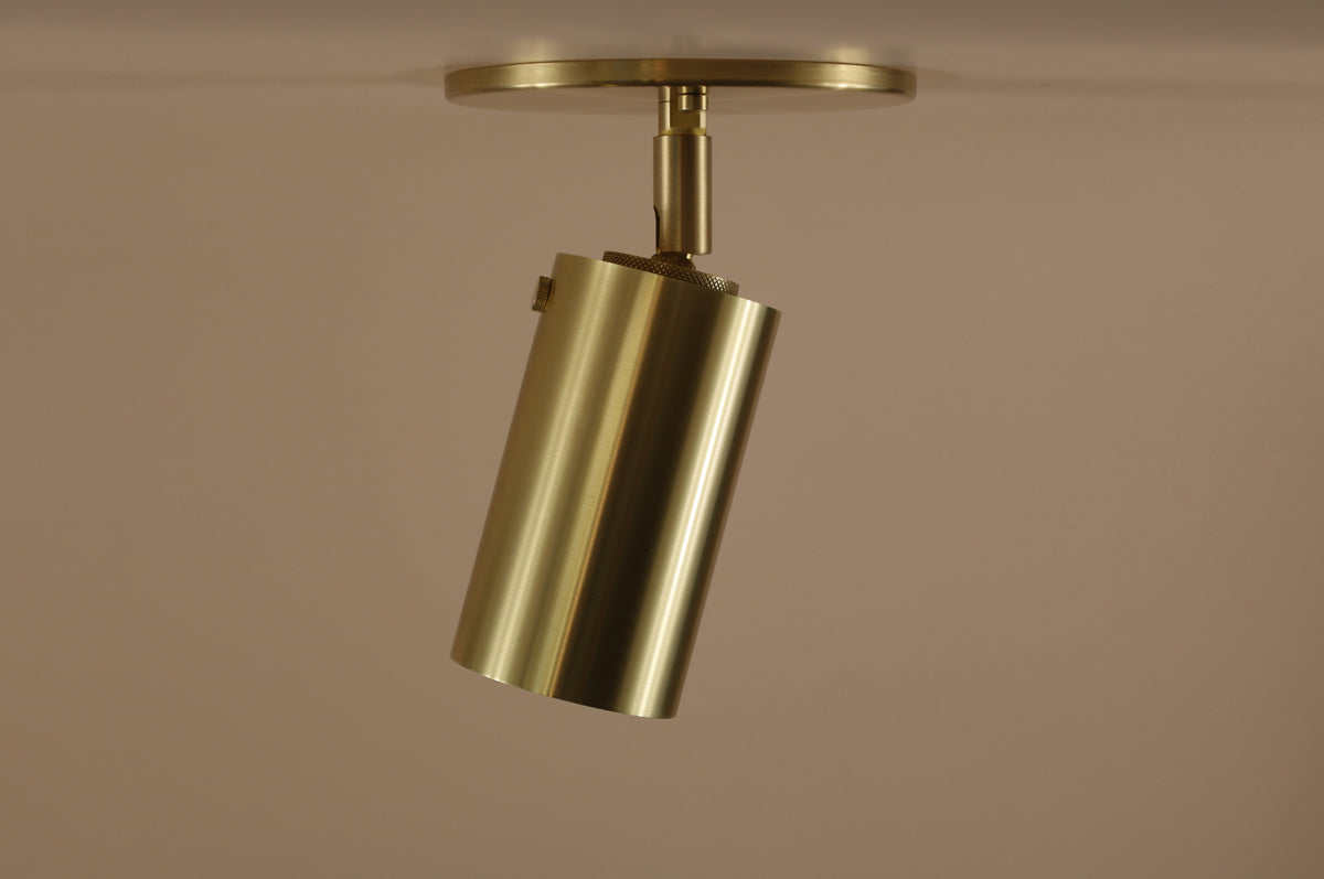 The Long Made Co. Brass Cylinder Adjustable Spot - Top Mount is a modern brass ceiling light with a cylindrical design, directional downlight, and circular base, shown against a plain beige background.