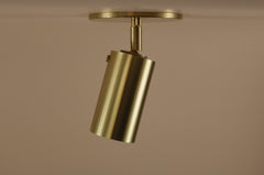 The Brass Cylinder Adjustable Spot - Top Mount by Long Made Co. features a cylindrical brass shade on a round base, shown against a beige background, offering classic mid-century modern style as an elegant adjustable downlight.